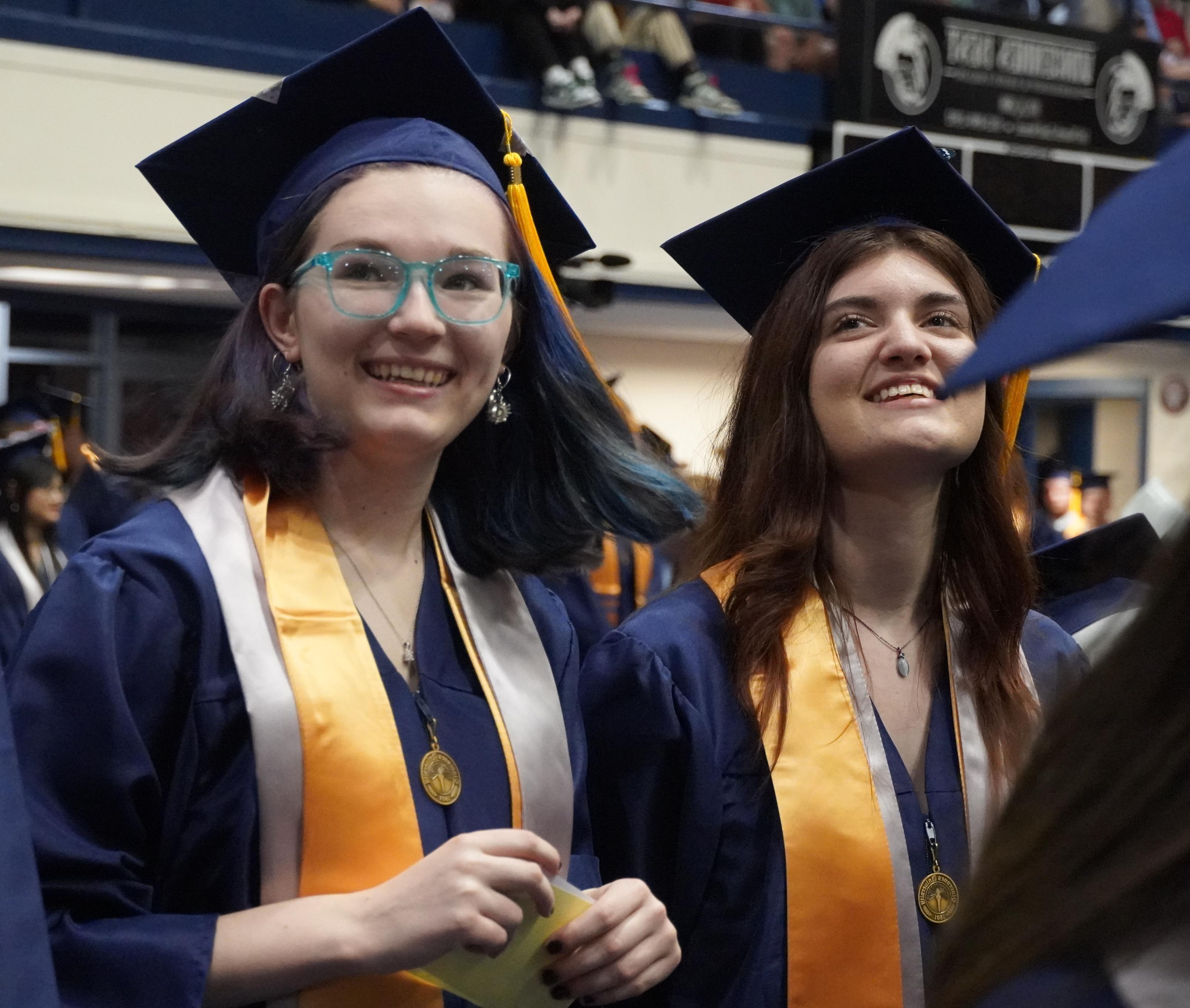 2个面带微笑的女毕业生戴着帽子和长袍. 一个戴着眼镜，头发颜色鲜艳.